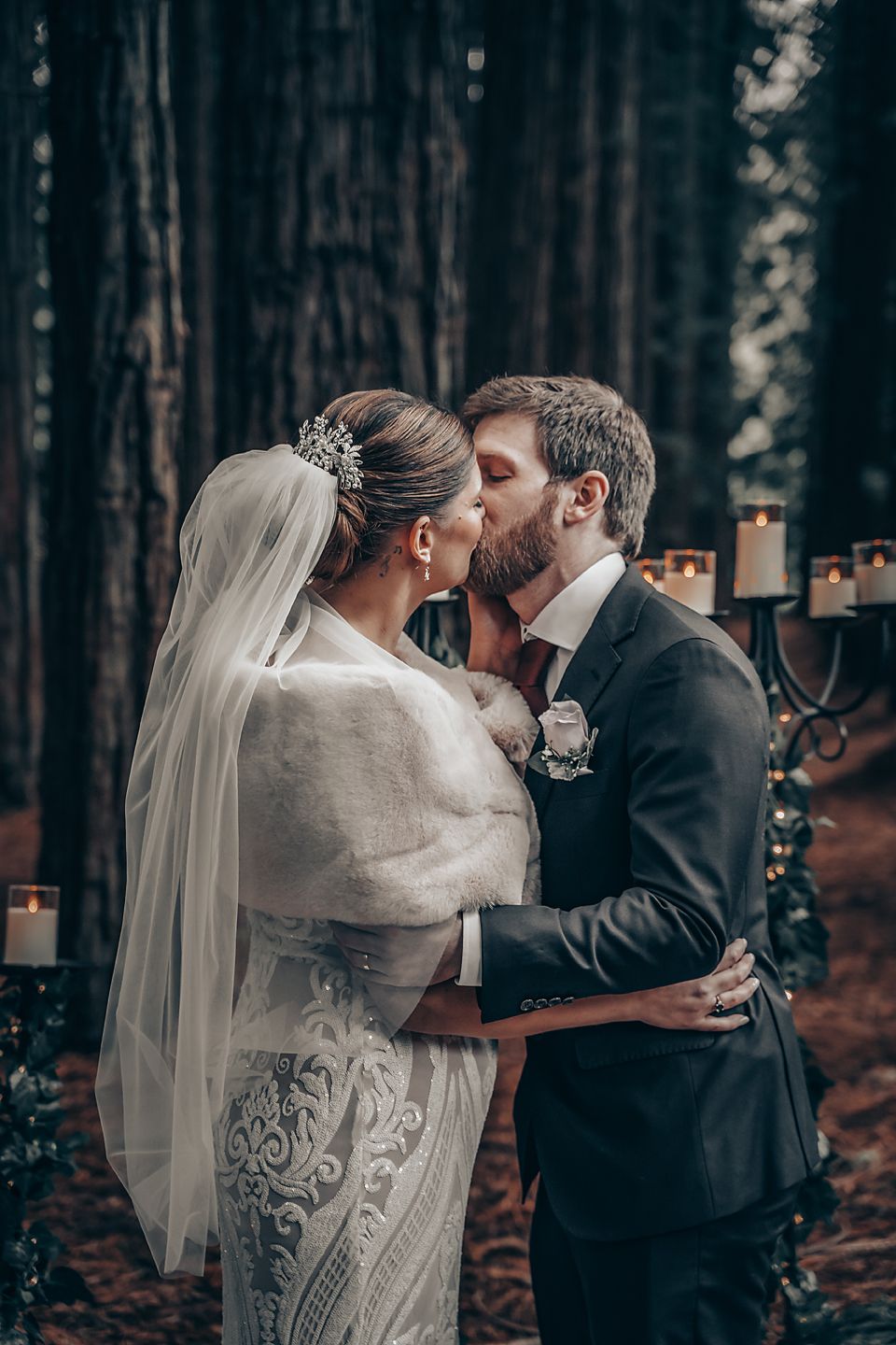 A romantic winter wedding moment in the forest with the bride in a fur shawl and veil kissing the groom among candles.