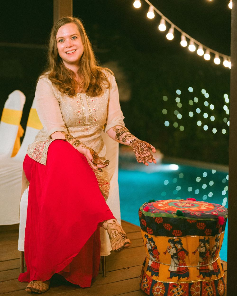 Person in a red dress and gold top sitting at an outdoor party with string lights.