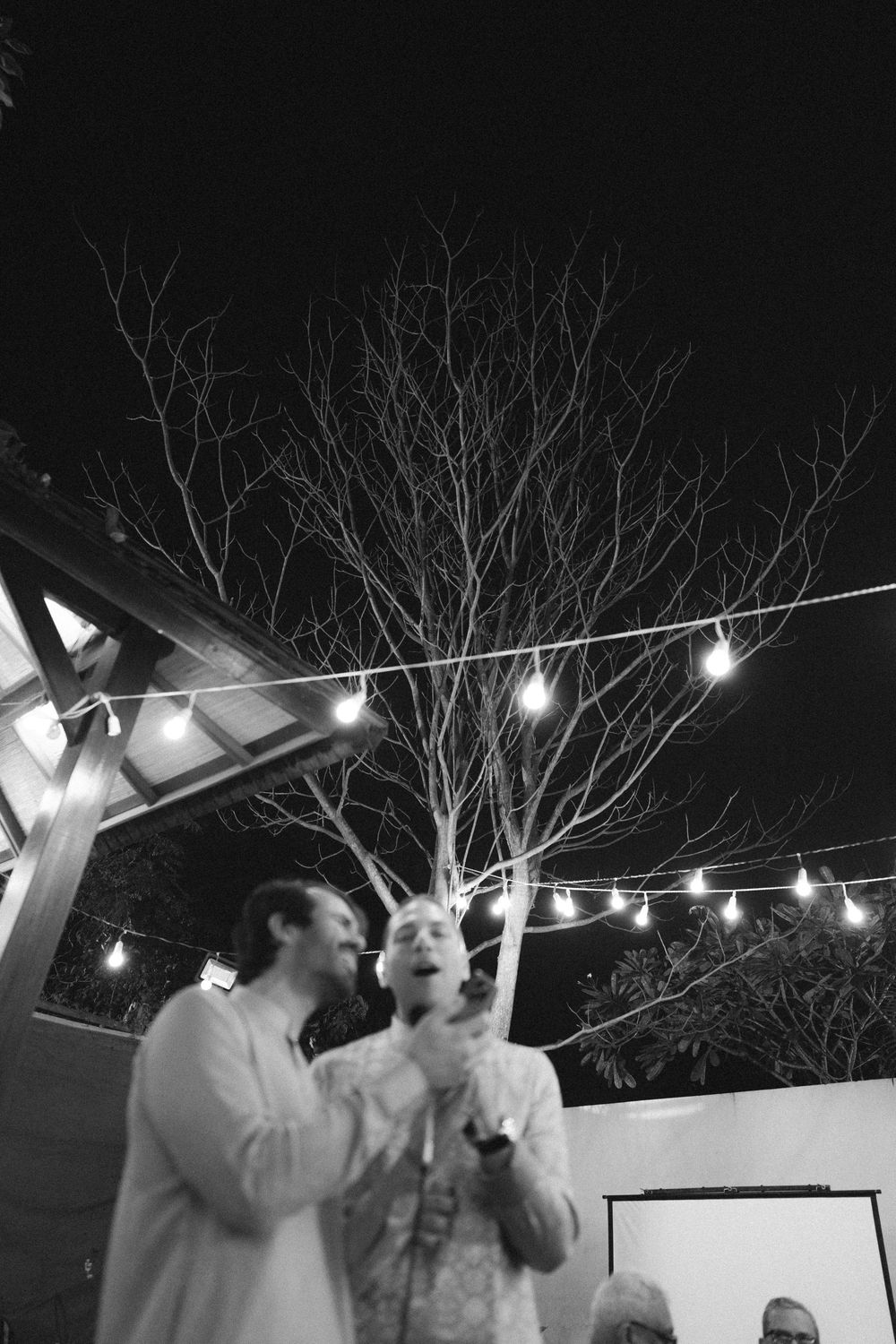 People singing and dancing under string lights at an outdoor nighttime celebration.