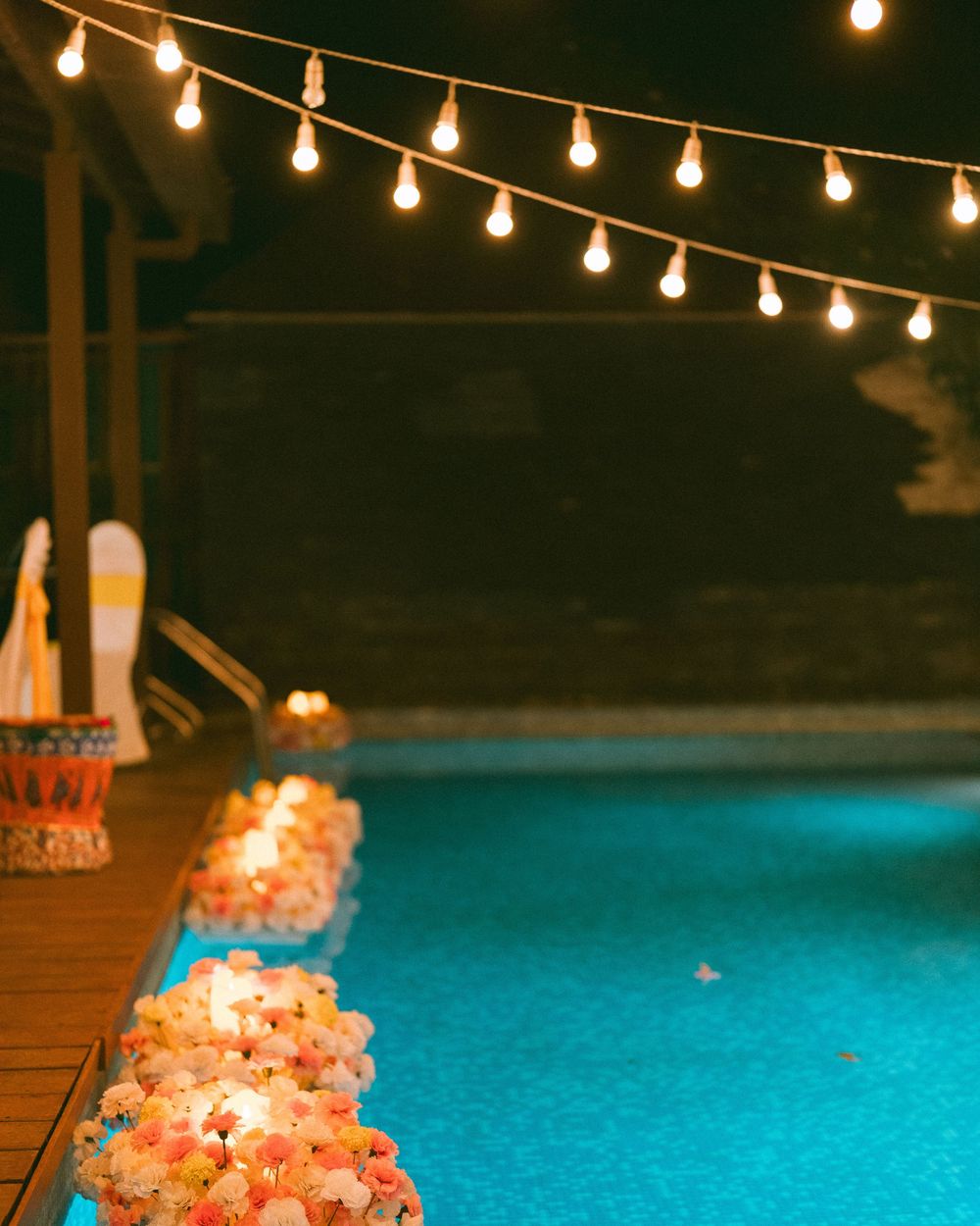 String lights illuminate a poolside party setting at night with decorative elements.