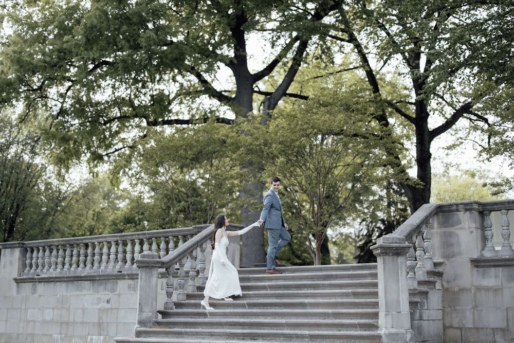 A couple walks down grand stone steps surrounded by trees and ornate balustrades in a formal garden setting.