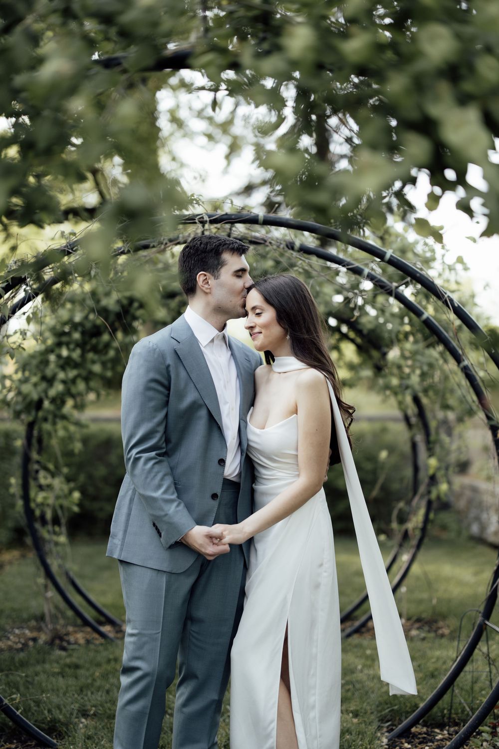 Romantic wedding couple poses intimately in an outdoor garden setting with a decorative arch.