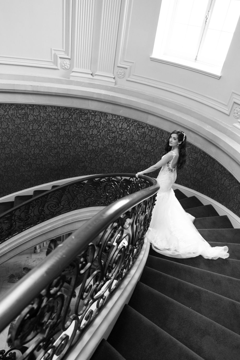 Bride in flowing white gown descends elegant spiral staircase with ornate black iron railings.