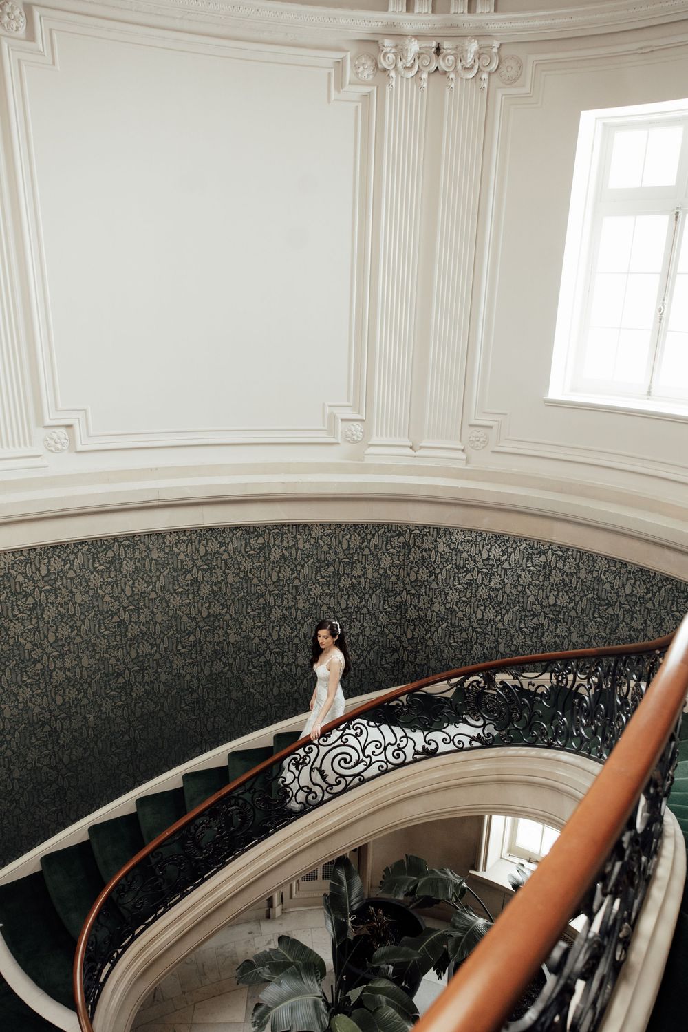 A dramatic spiral staircase in an elegant white interior features ornate baroque detailing and a hanging chandelier.