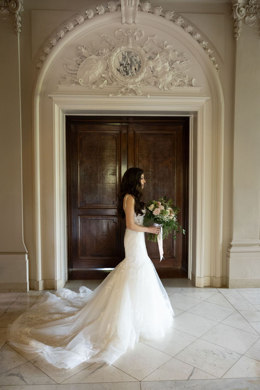 An elegant room with arched doorways and ornate crown molding features a flowing white wedding gown against dark wood panels.