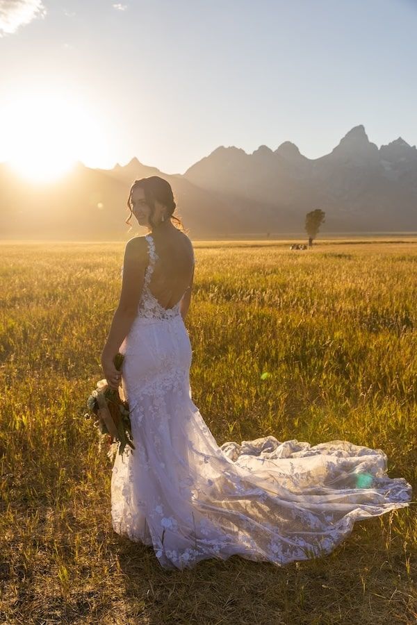A bride in a flowing white dress sits in a golden field at sunset with majestic mountain peaks silhouetted in the background.