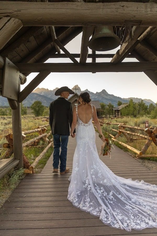A couple walking on a wooden bridge with mountains in background as a white lace wedding dress train trails behind them.