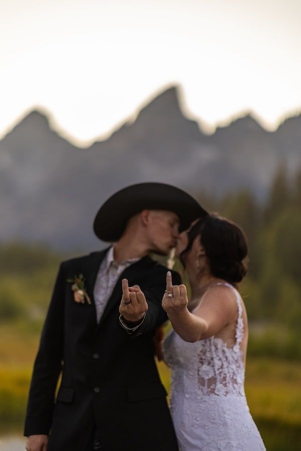 A bride and groom share a tender moment in an outdoor setting with mountains and soft sunset lighting in the background.