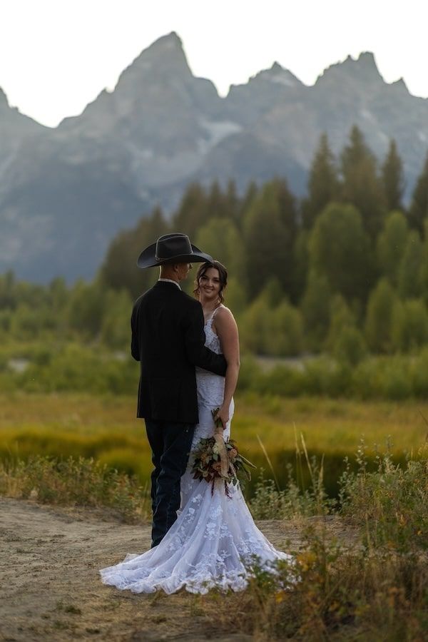 A couple in wedding attire embraces while standing in a meadow with majestic mountains rising in the background.