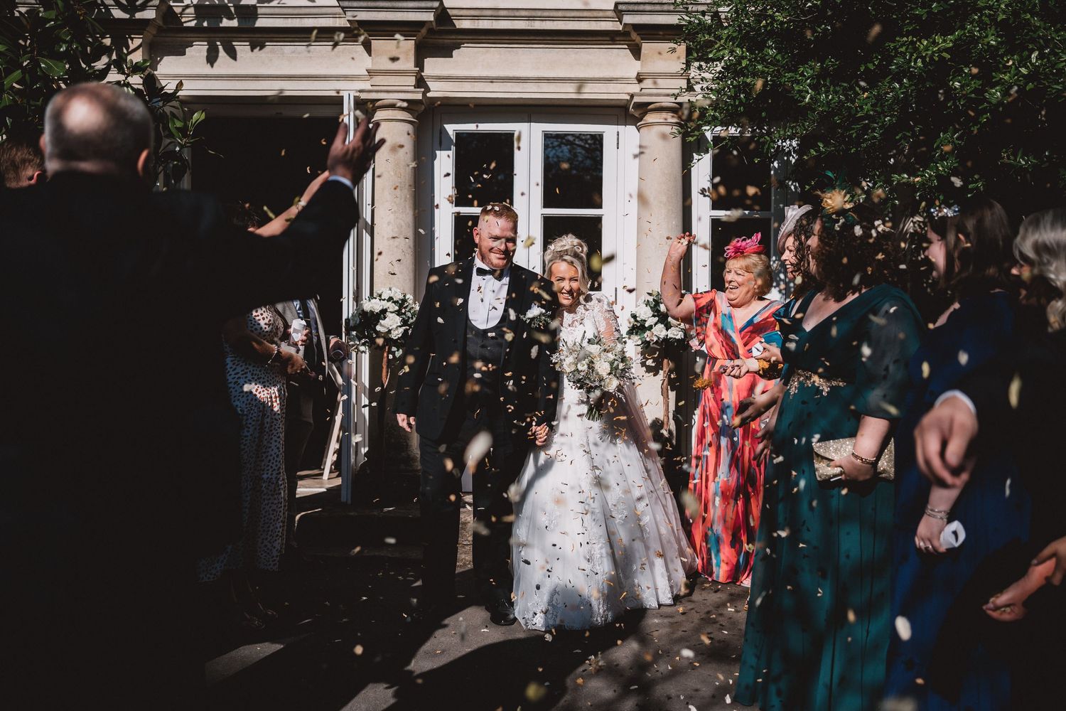 Wedding party exits venue while guests throw confetti during joyful celebration at elegant building entrance.