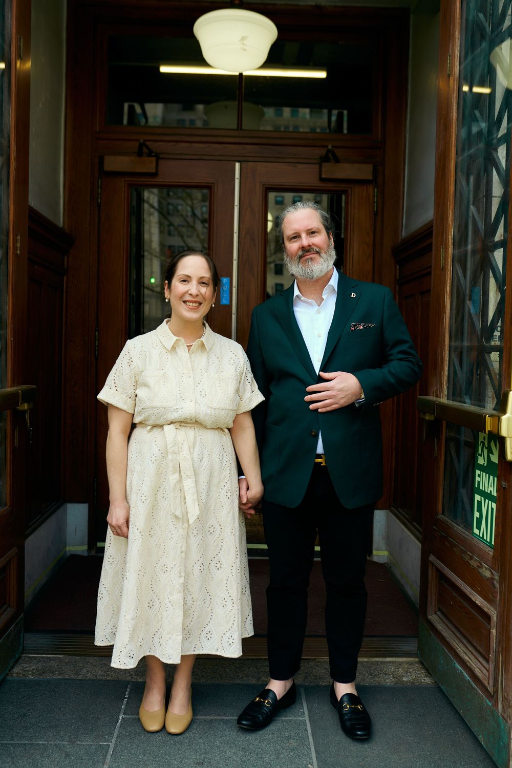 Two people stand in an entryway, one wearing a cream dress and another in a dark green blazer. Wedding photography