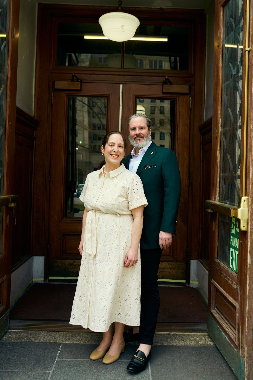 A couple poses in formal attire in front of a wooden doorway, the woman in a cream dress and the man in a dark jacket. Wedding photography