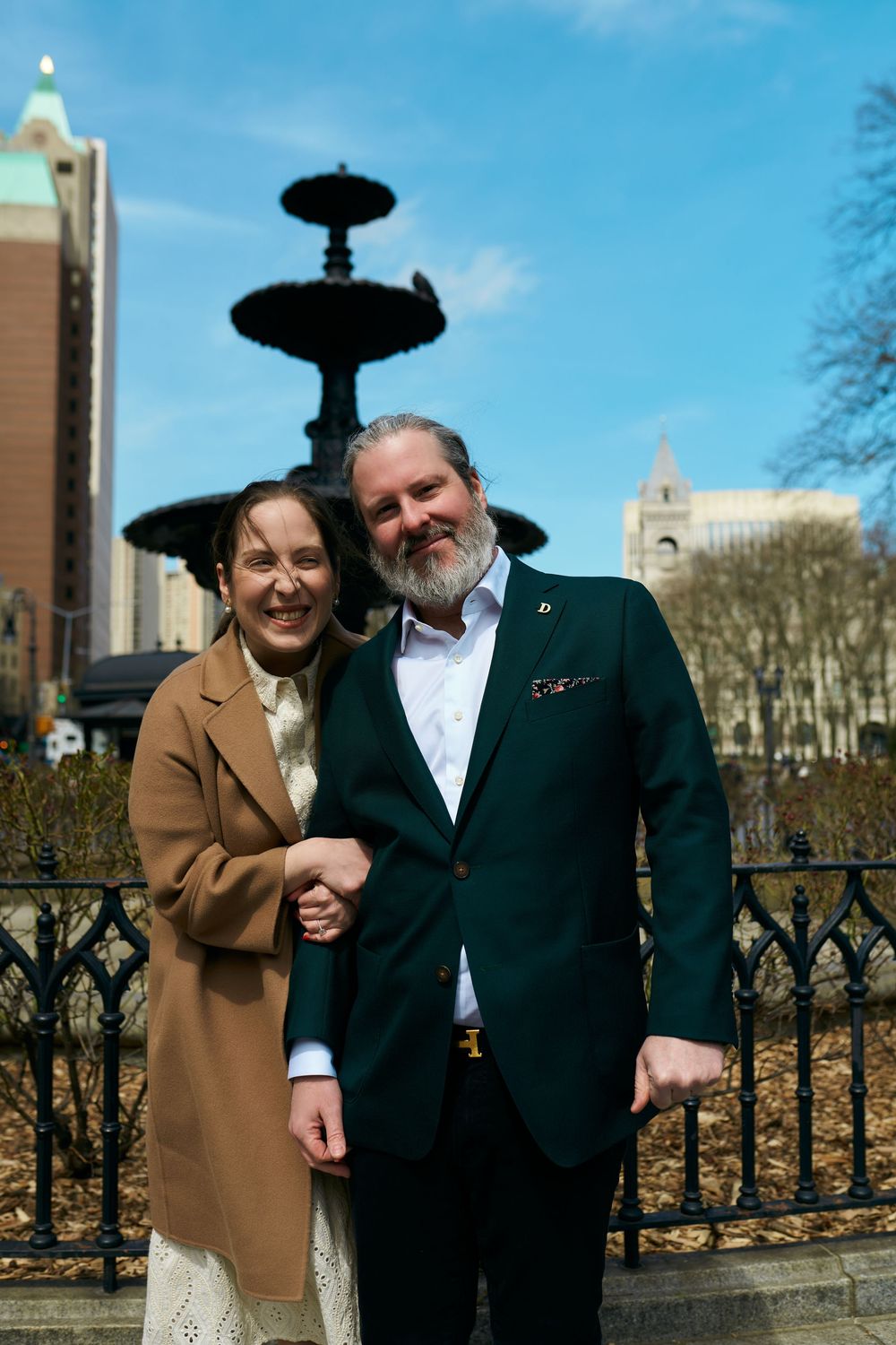 A couple poses together near an ornate fountain in an urban setting on a bright day. Wedding photography