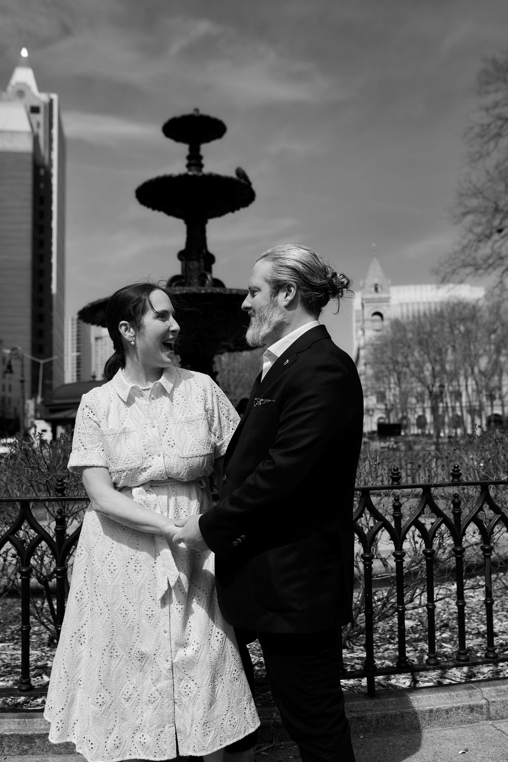 A couple in vintage-style clothing share a romantic moment by an ornate fountain in a city park on a cloudy day. Wedding photography