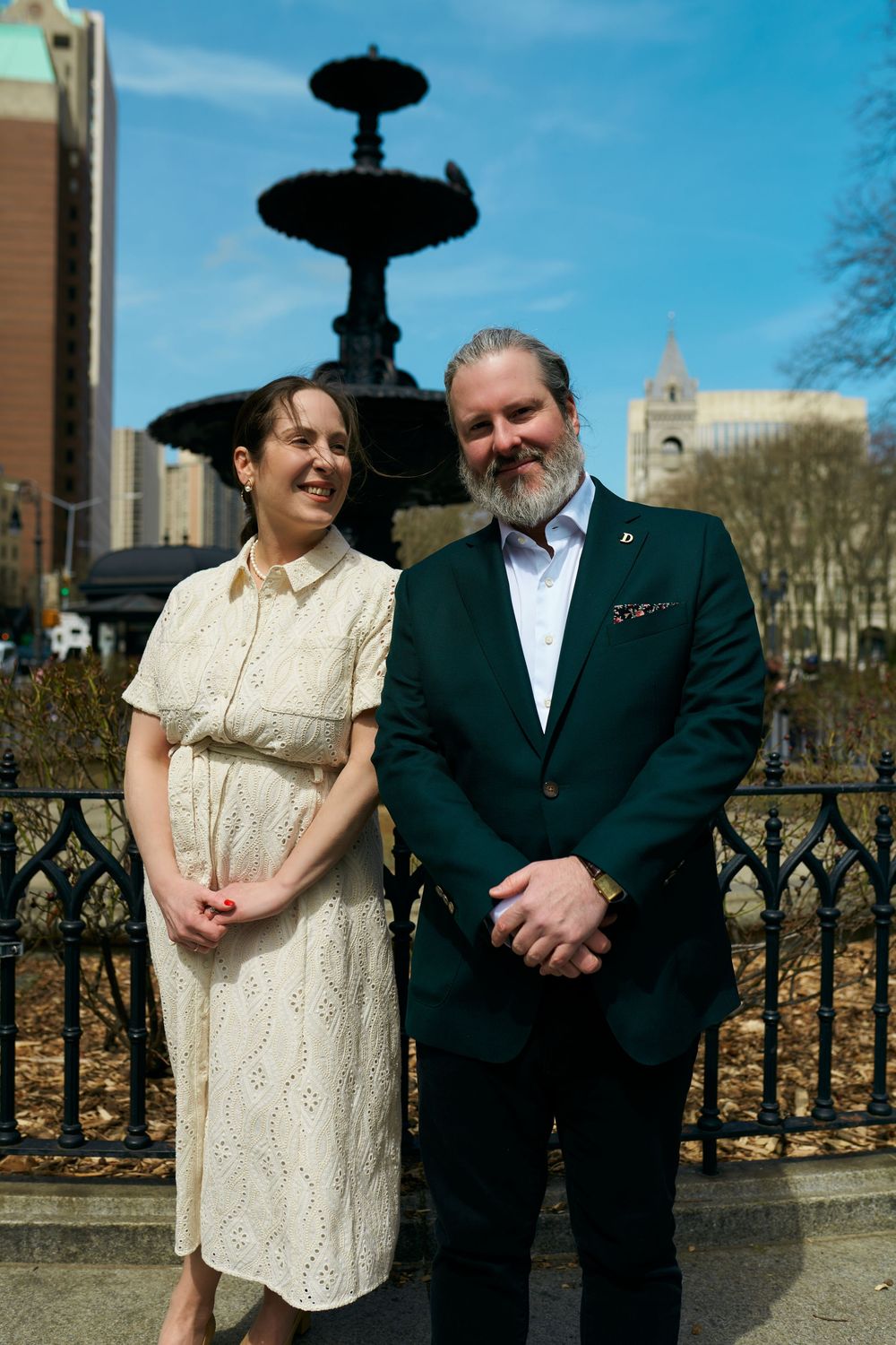 Well-dressed couple poses in front of ornate fountain in urban park setting on sunny day. Wedding photography