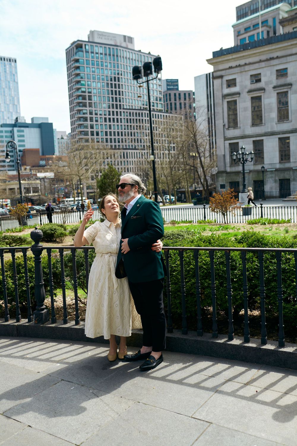 A couple embraces on a sidewalk with city buildings and greenery in the background on a sunny spring day. Wedding photography