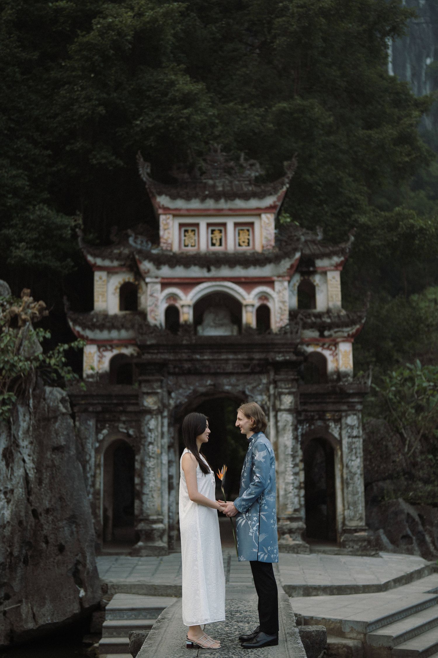 A couple stands together in front of an ornate traditional Asian temple gateway with decorative architecture.