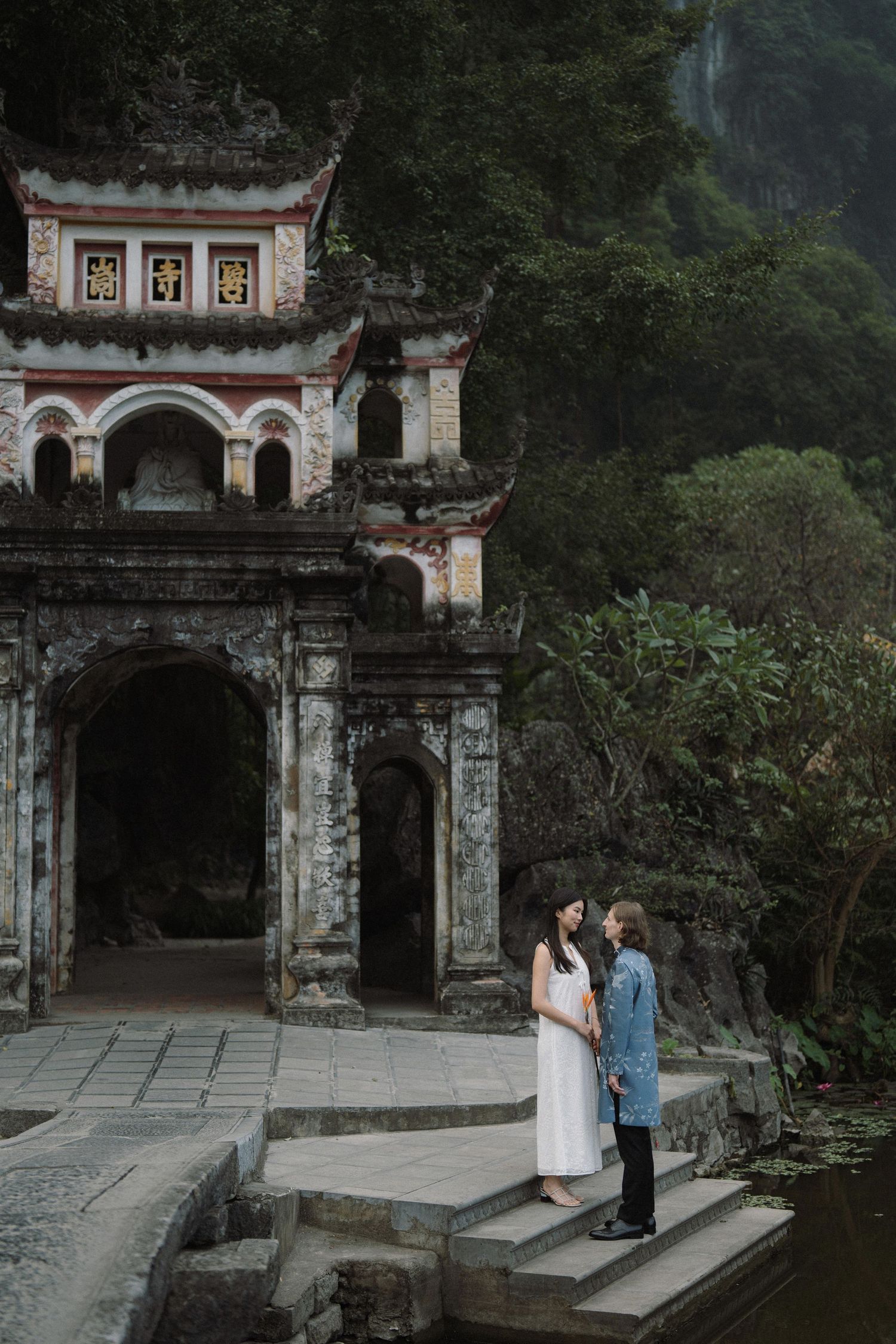 A couple stands on stone steps before an ancient temple entrance with Chinese architectural details.