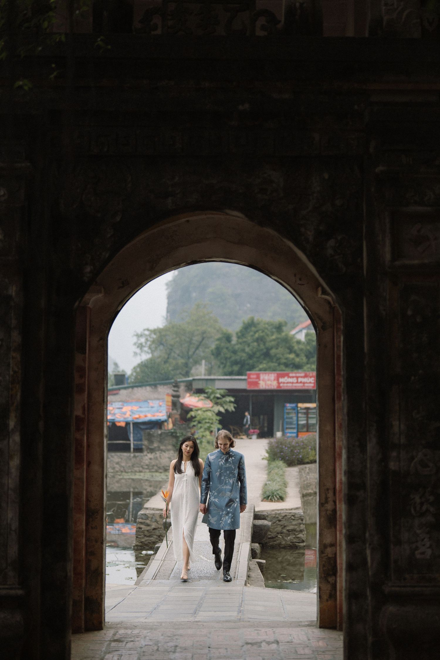 Silhouetted couple seen walking through stone archway on path during outdoor wedding photoshoot.