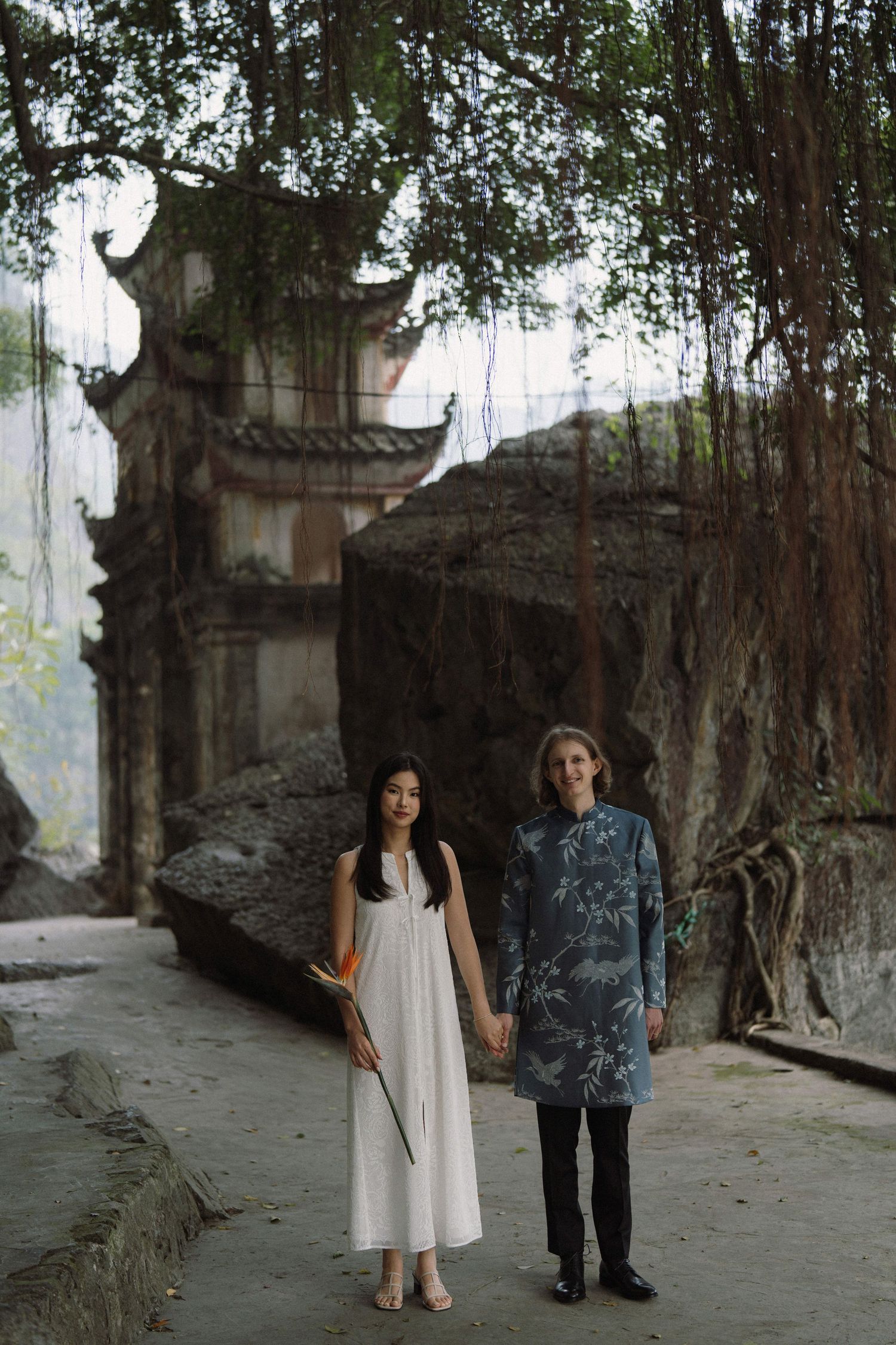 Couple walking along a stone path surrounded by ancient temple ruins and lush foliage.