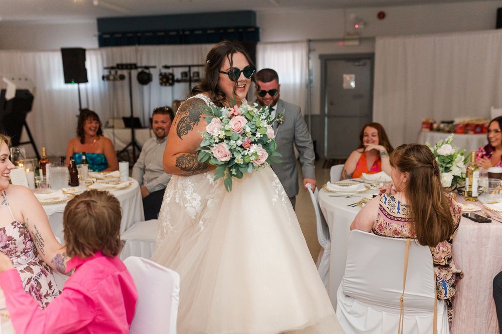 A wedding reception scene in a white banquet hall with guests seated at decorated tables and a bride holding a bouquet.