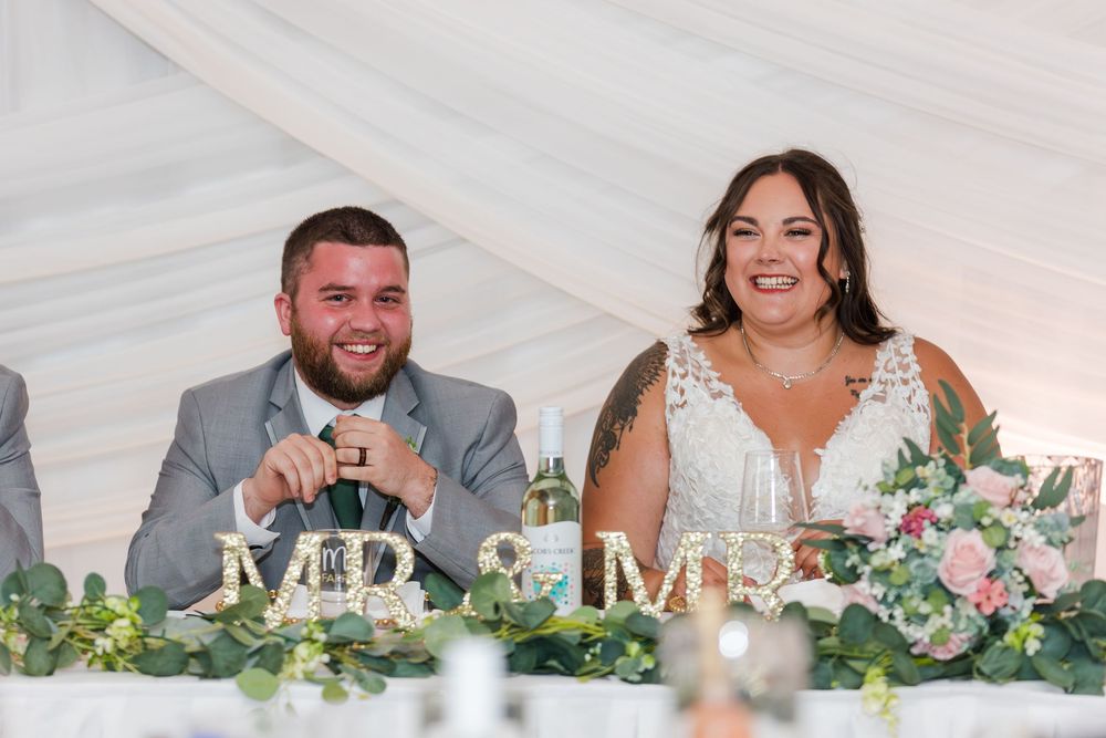 A couple shares a joyful moment at their wedding reception head table decorated with fresh greenery and flowers.