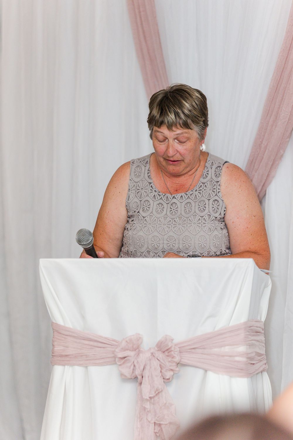 Someone in a gray lace dress speaks at a wedding podium draped with white and pink fabric decorations.