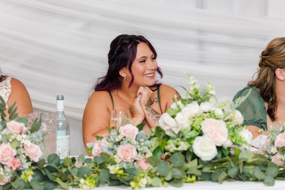Someone at a wedding reception table laughs while sitting behind elegant white floral centerpieces and greenery.