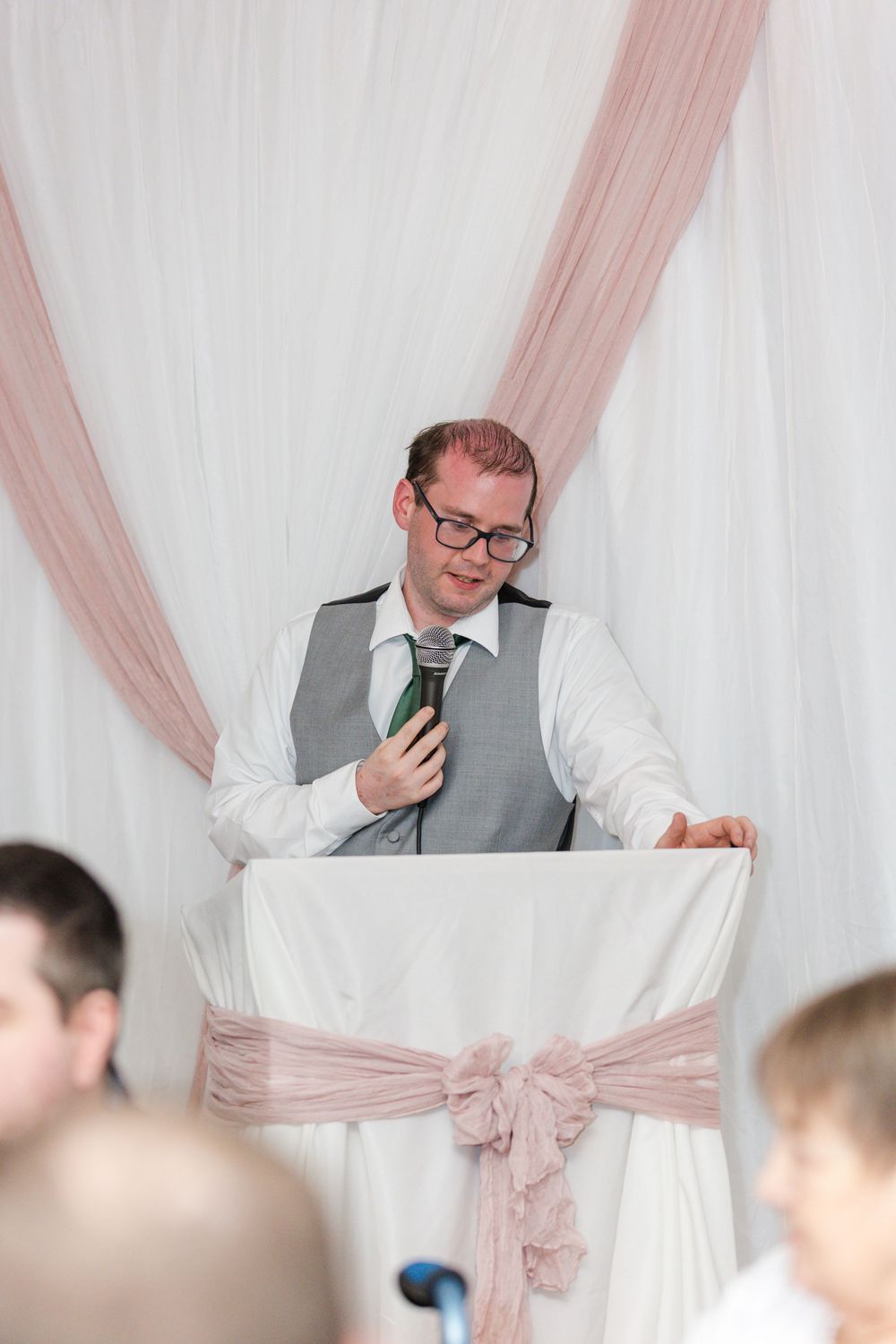 Man in gray vest and glasses gives emotional wedding reception speech while holding paper notes.