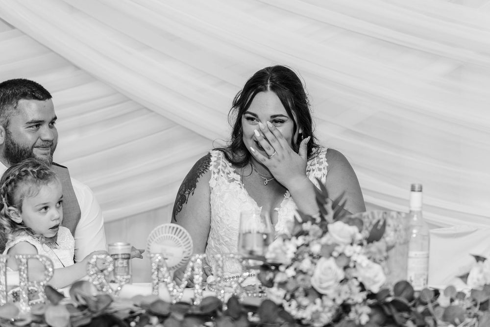 Wedding reception guests at a head table share a joyful moment surrounded by floral decorations in a white tent.