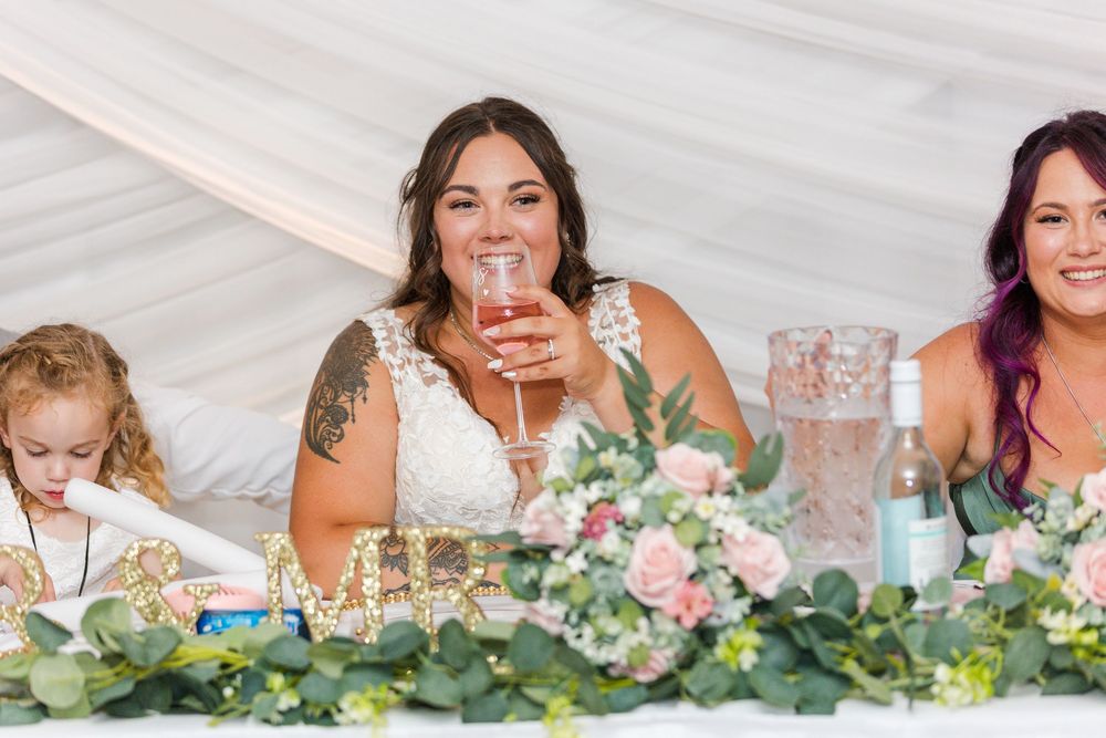 Wedding party seated at decorated head table with floral arrangements and white tent draping.