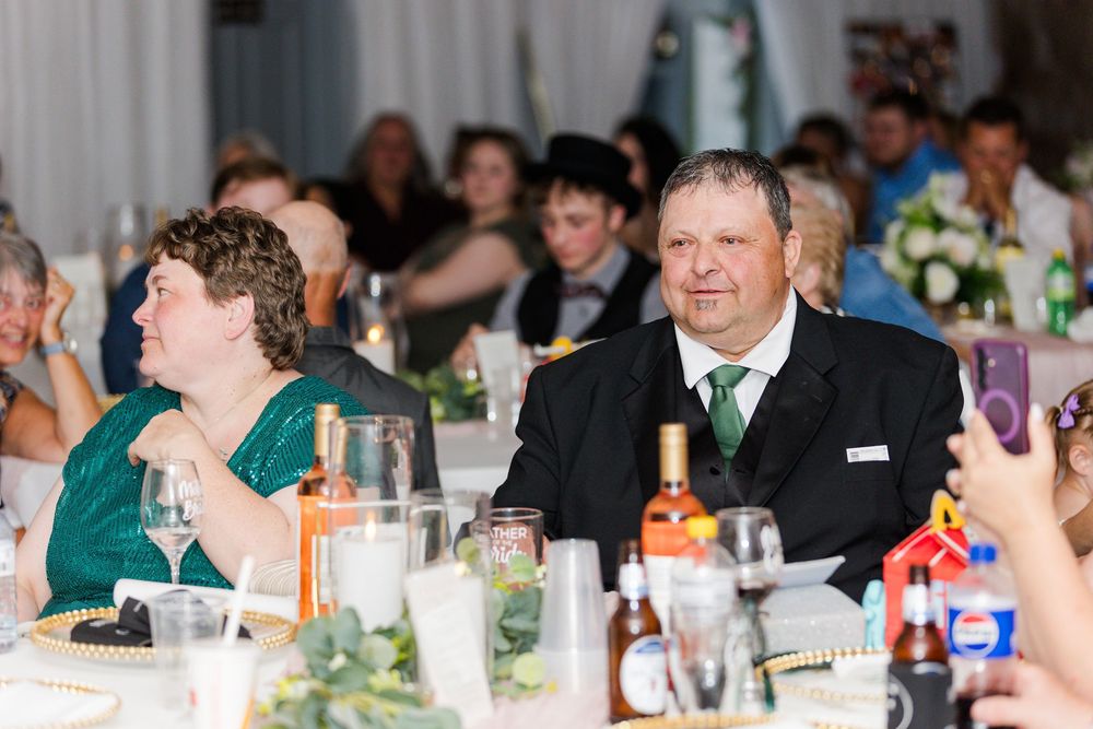 Wedding guests seated at a reception table smile during dinner service with wine glasses and drinks.