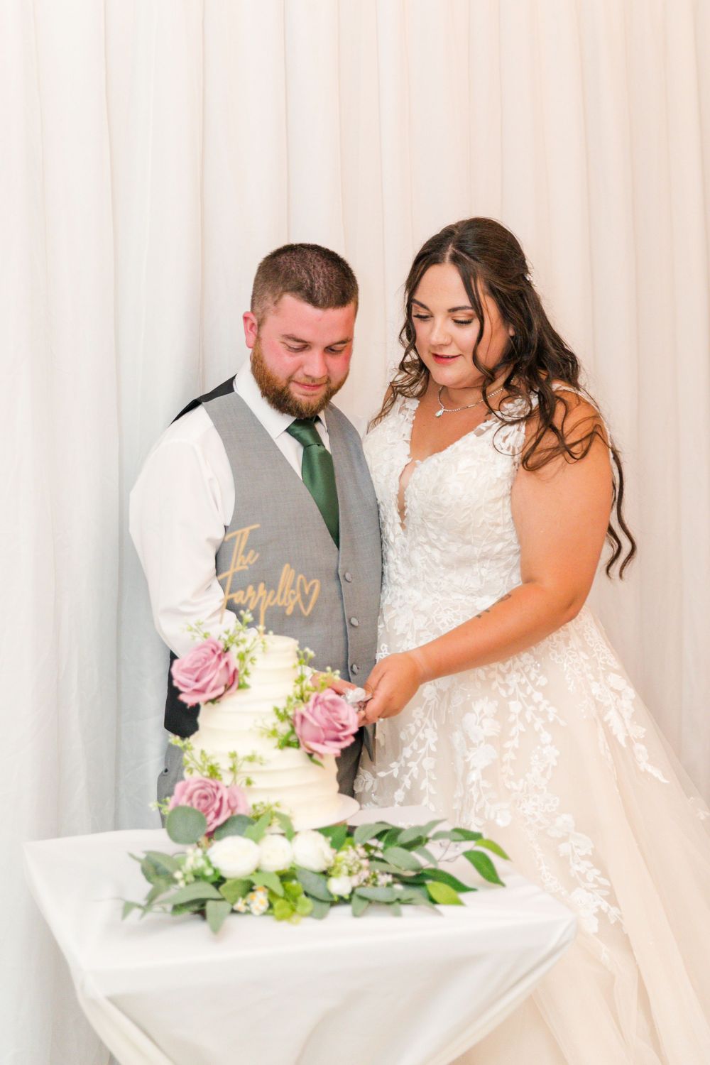A couple in wedding attire interacts with their wedding cake at the reception with white curtain backdrop.