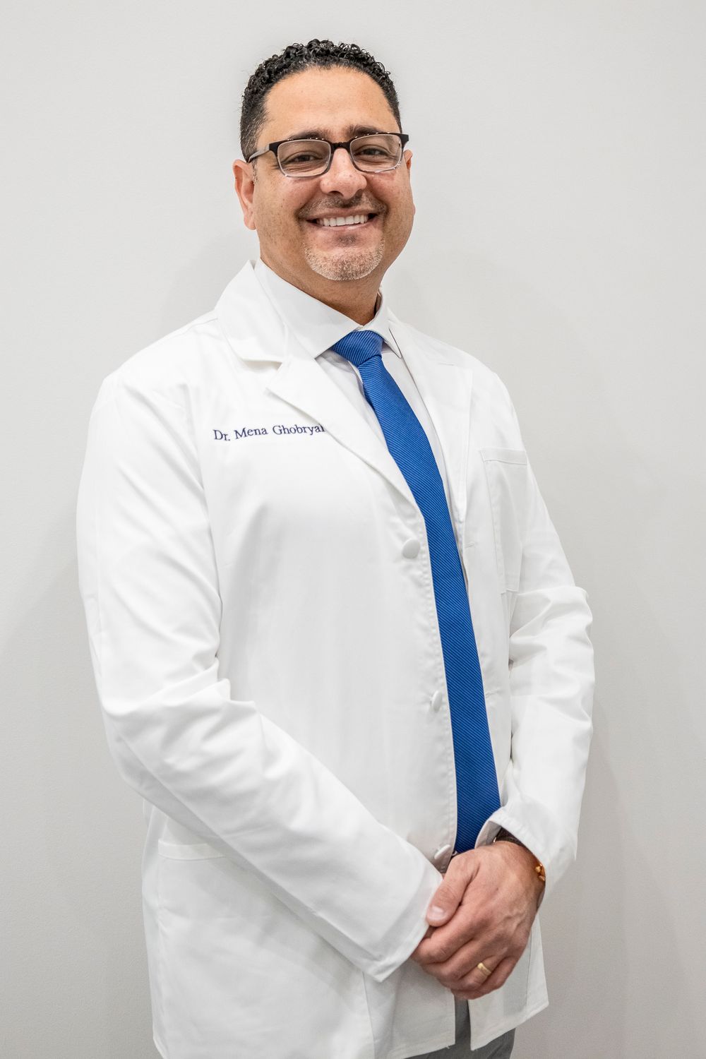 Healthcare professional in white coat with blue tie poses against light background.