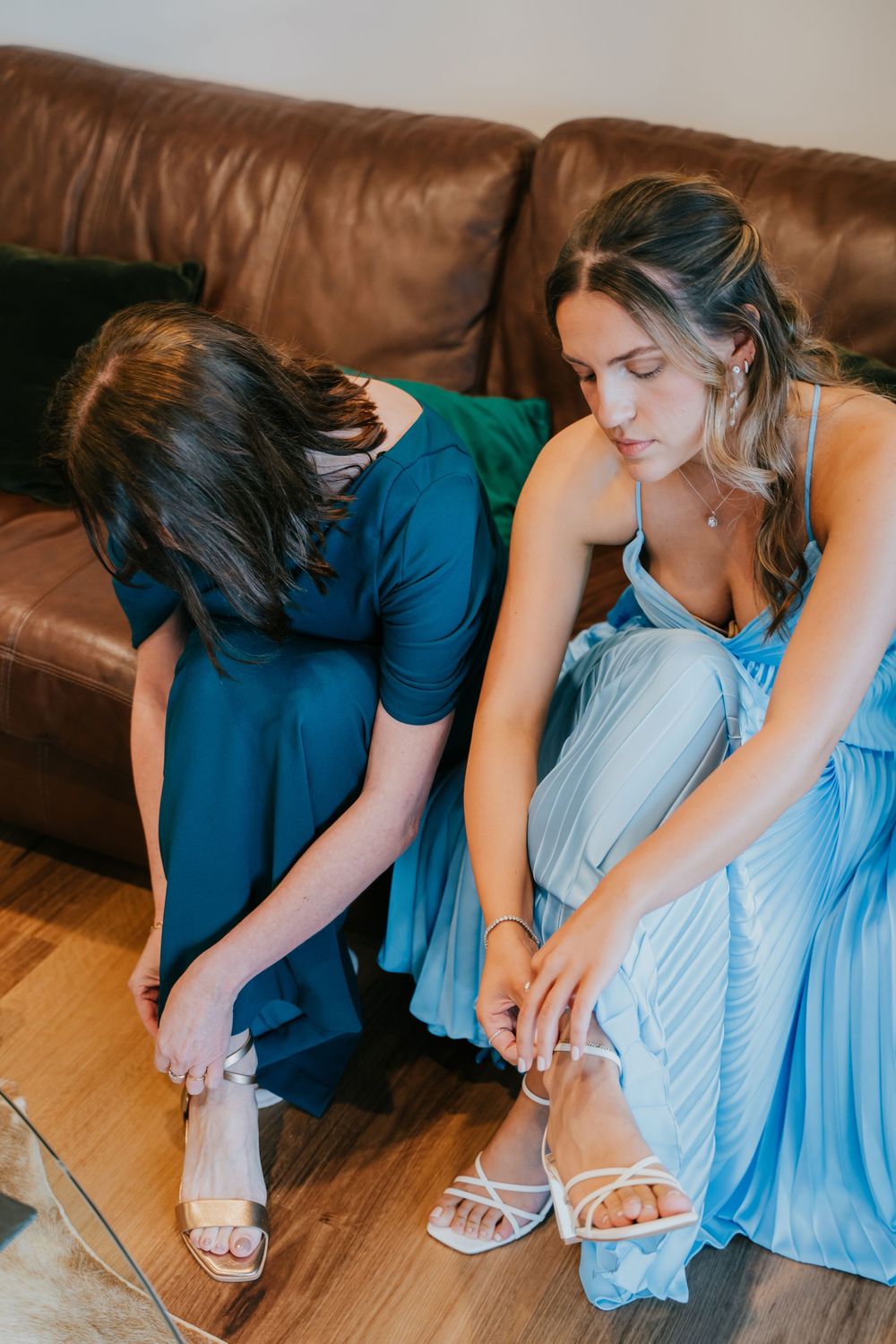 Someone in a blue dress sits on hardwood floor putting on white sandals and jewelry.
