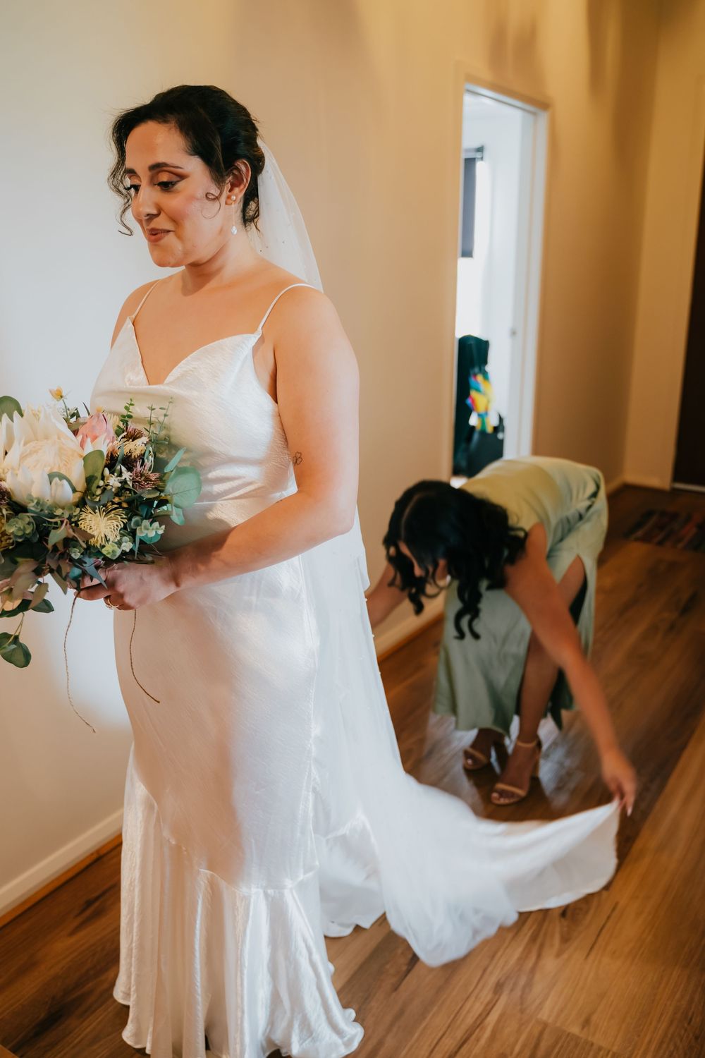 A bride in a flowing white gown and veil holds a colorful bouquet while getting ready in a sunlit room.