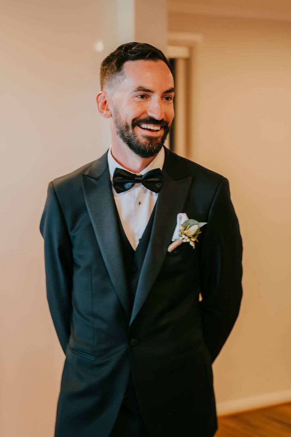 A beaming groom in a classic black tuxedo and bow tie with a boutonniere smiles during wedding preparations.
