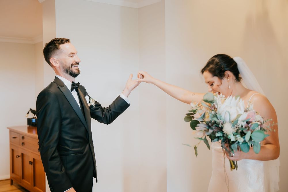 Newlyweds share joyful dancing moment together in elegant indoor setting with floral bouquet and natural lighting.