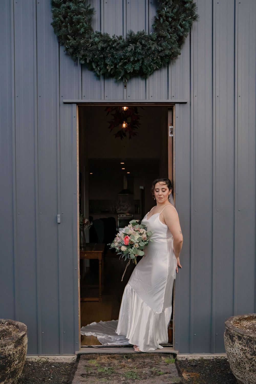 Someone in a white wedding dress stands in a doorway at a grey building decorated with holiday wreaths.