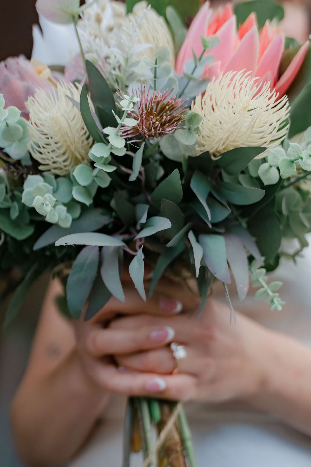 Close-up of hands holding a beautiful wedding bouquet with pink proteas, white chrysanthemums and eucalyptus leaves.