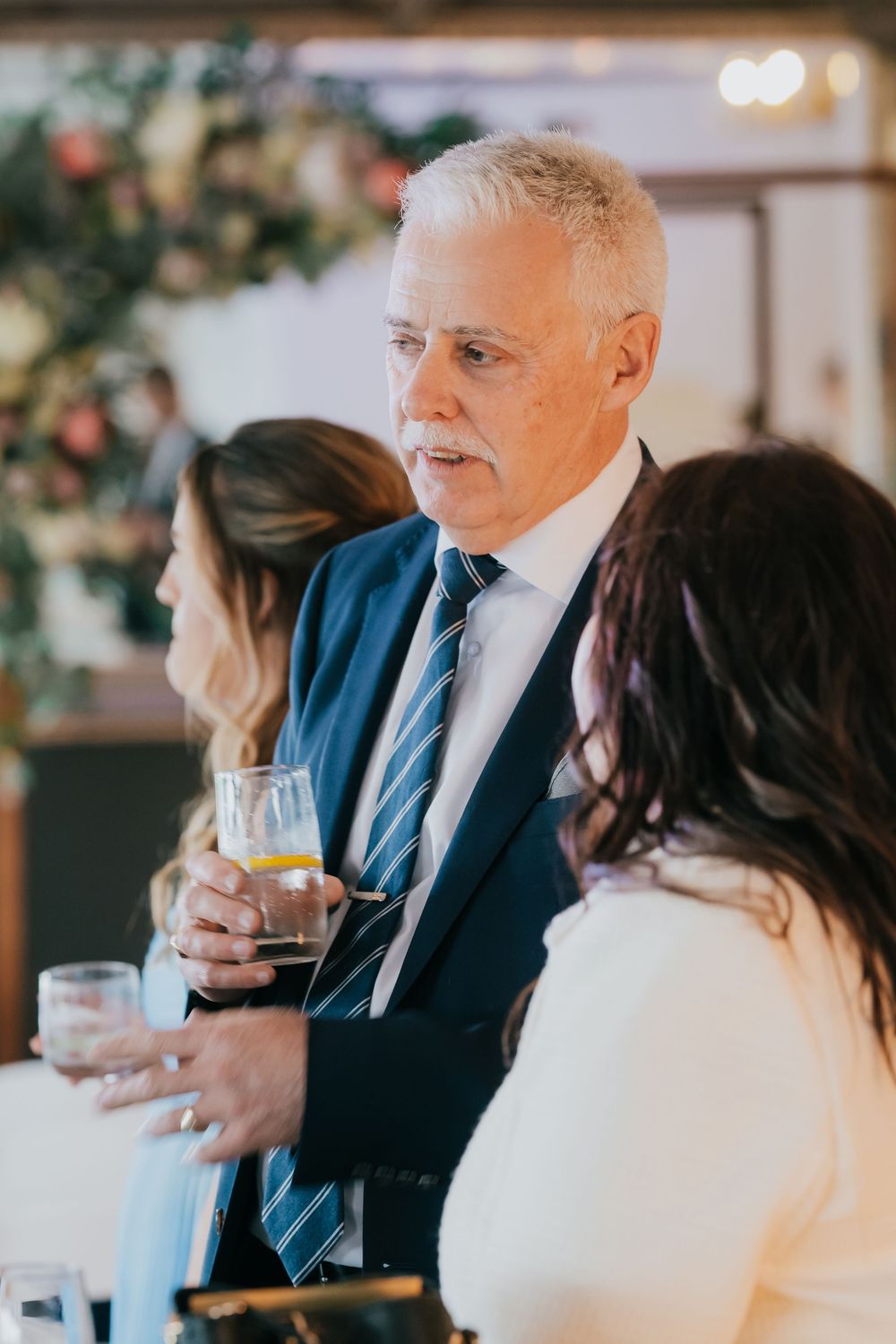 Well-dressed guests enjoying drinks and conversation at an elegant indoor celebration with floral decor.