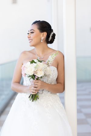 Bride in a white wedding gown holds a beautiful bouquet of blush and white flowers on a sunlit porch.