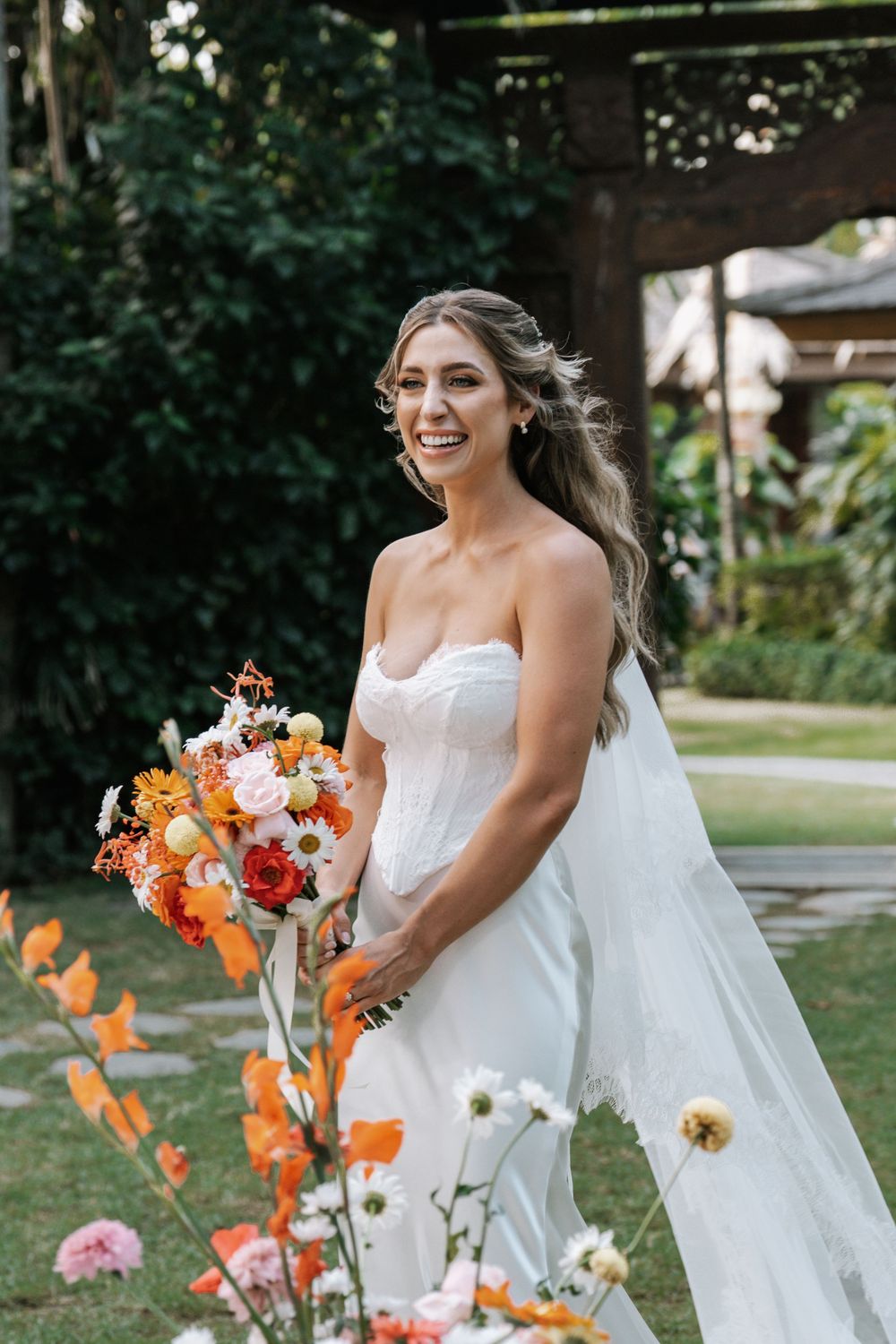 A bride in a strapless white gown holds a vibrant orange and pink bouquet while standing in a garden setting.