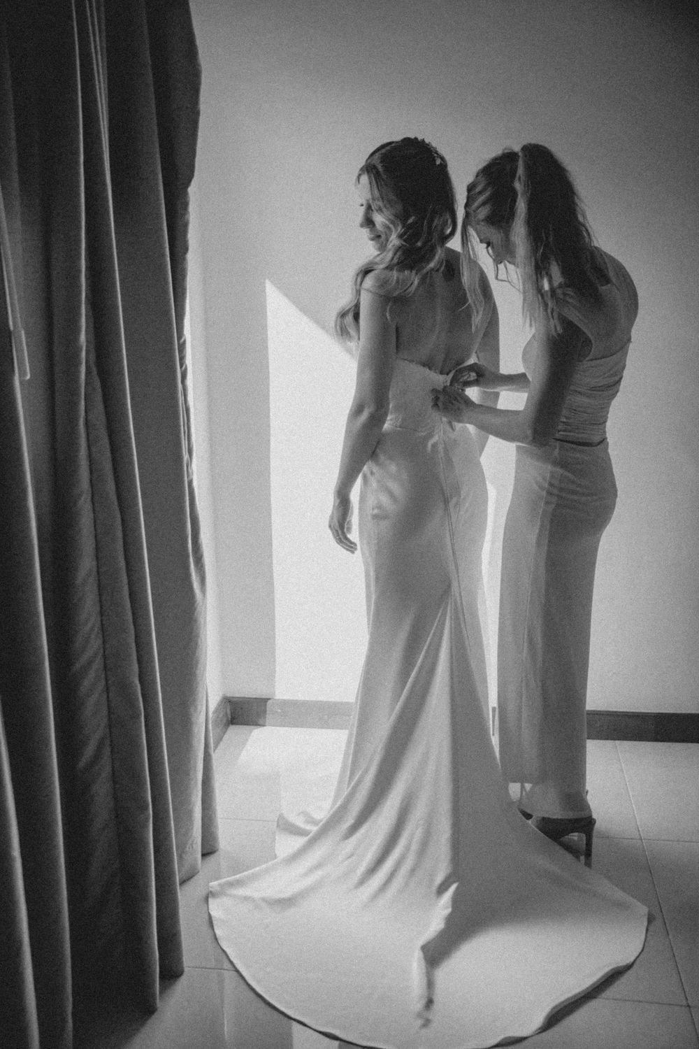 Artistic black and white shot of two people in white dresses conversing by a window with curtains.