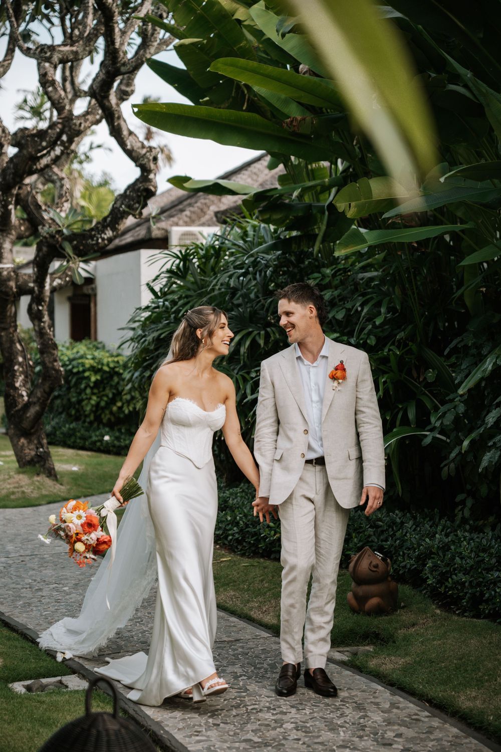 A couple in wedding attire walking together on a garden path surrounded by lush tropical greenery and trees.