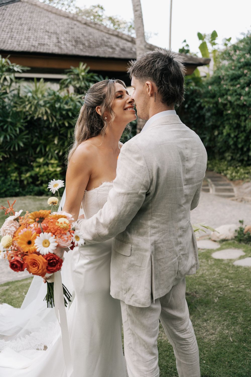 A romantic wedding moment captured in a tropical garden setting with bright orange and white floral bouquet.