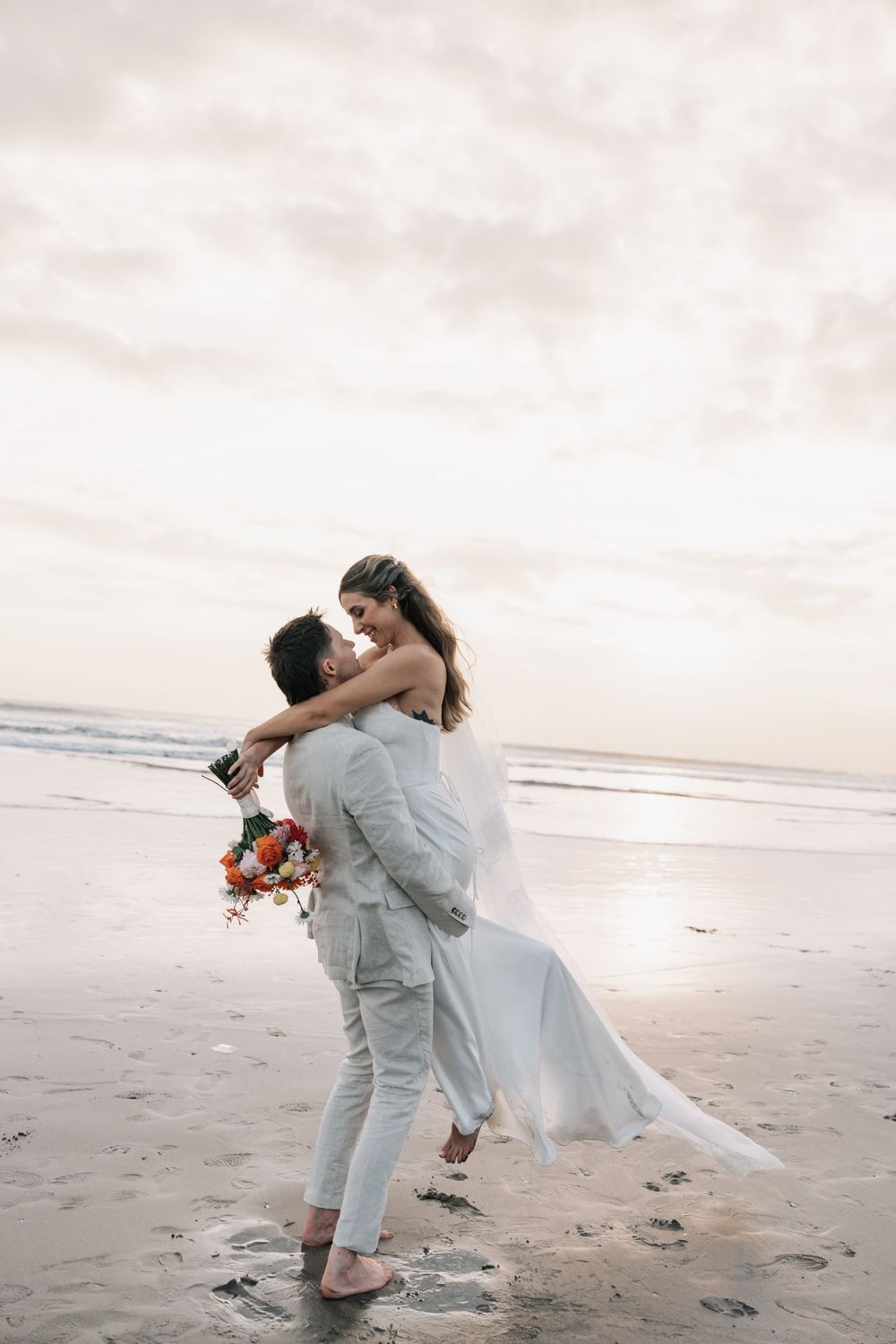 A romantic couple embraces during a sunset beach wedding photoshoot with the bride holding a floral bouquet.