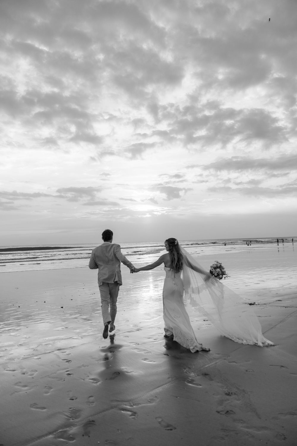 Black and white beach panorama showing a couple in formal attire walking together along the shoreline at sunset.
