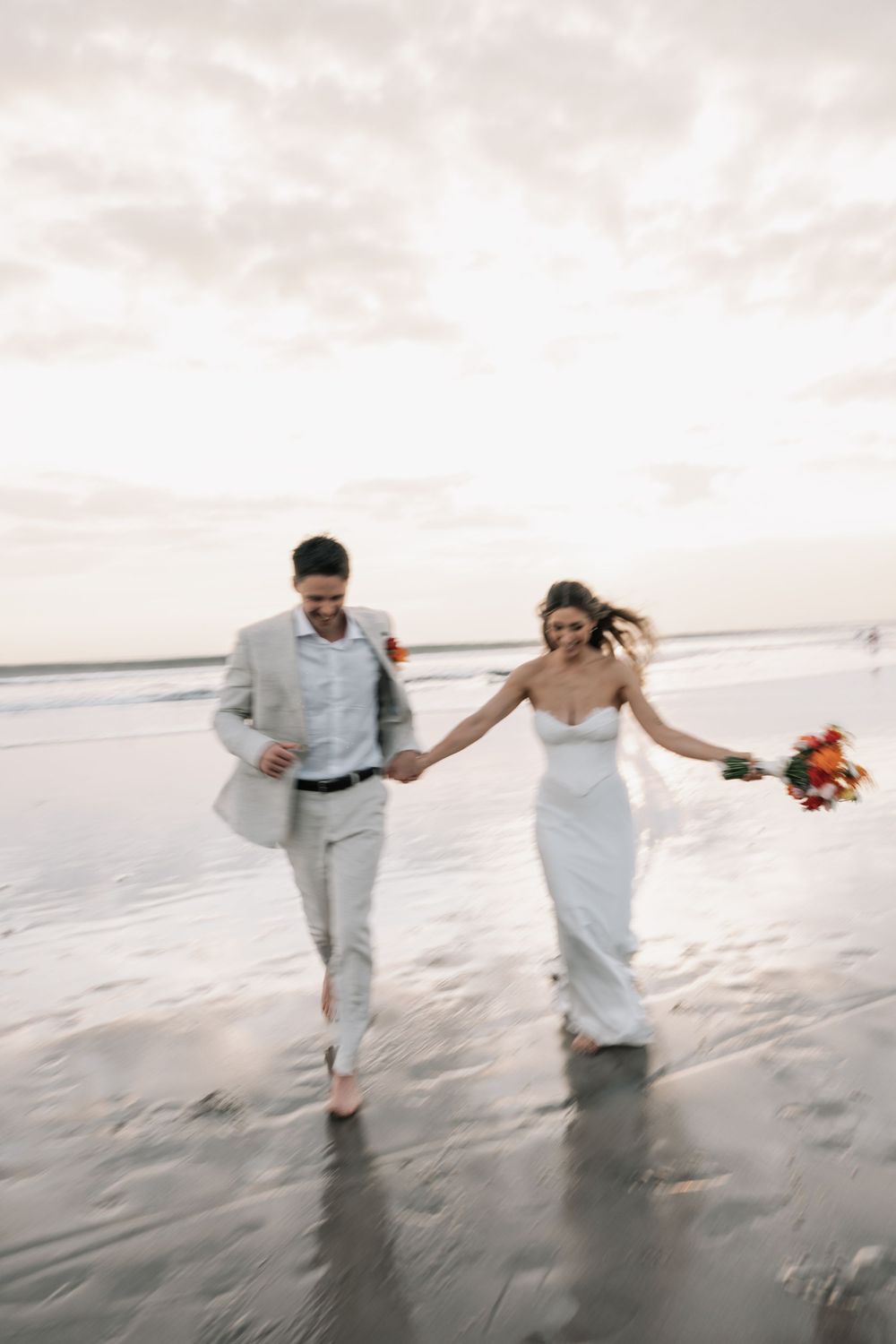 A newlywed couple in white attire walks joyfully along a misty beach at sunset during their wedding photo session.