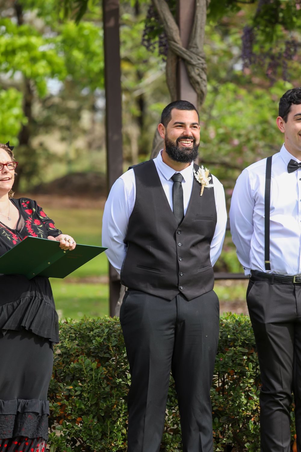 Groomsmen in formal attire pose outdoors in front of green hedges during a wedding ceremony.